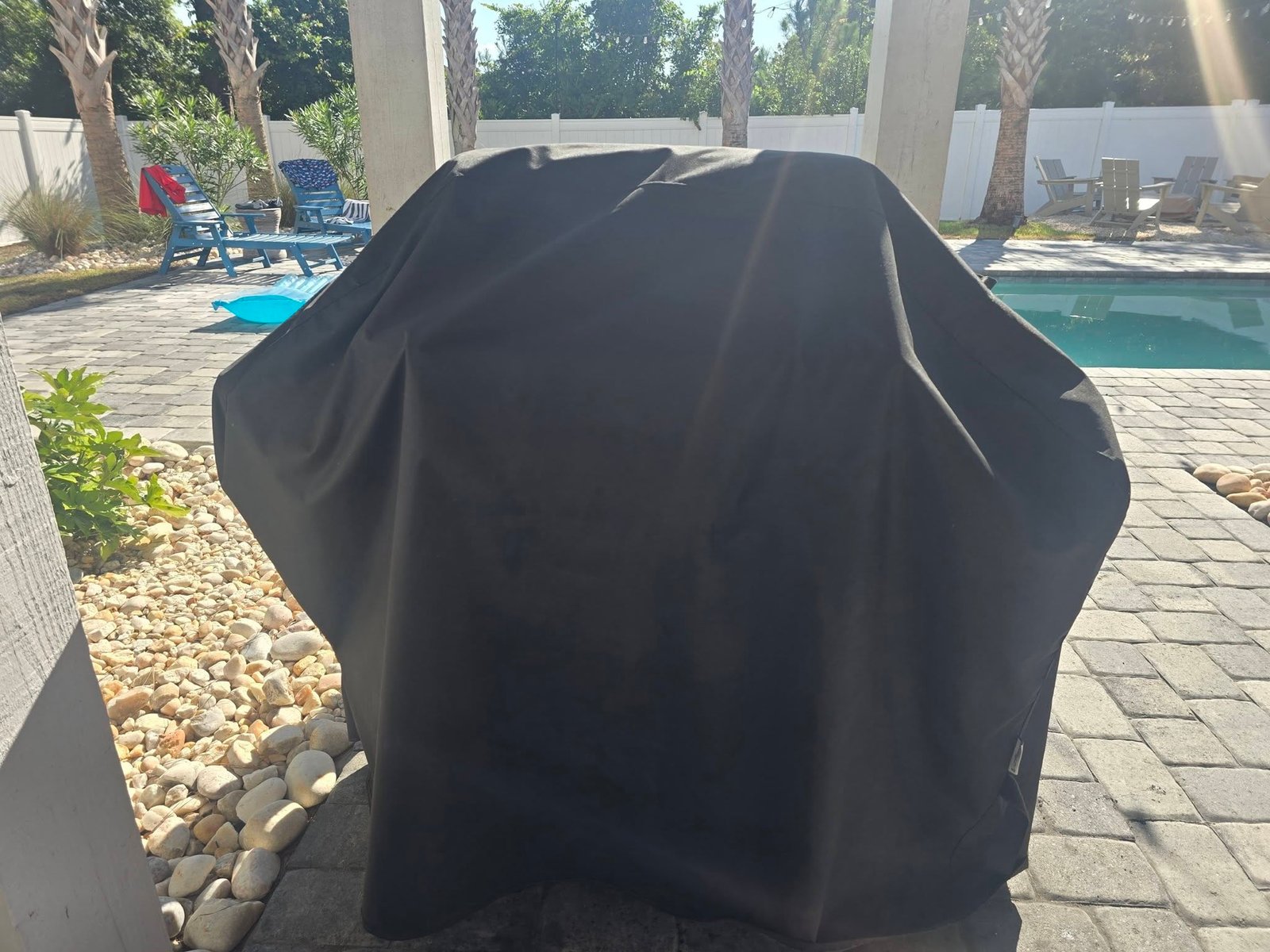 A backyard grill under a fitted cover beside a pool, palm trees behind